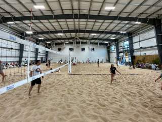 Indoor sand volleyball facility with multiple courts inside a large metal building, where players are actively competing on a sandy surface, divided by nets, with spectators seated along the sides under bright overhead lights.