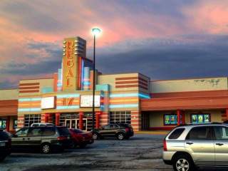 Exterior view of a Regal movie theater with a retro-style facade featuring neon accents and a vertical ‘Regal’ sign, with a parking lot and several cars in the foreground under a colorful sunset sky.