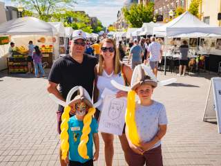 A family poses in the middle of a street lined with art vendor tents during the Carmel International Arts Festival. Two children wear balloon Viking helmets while crowds browse booths in the background.