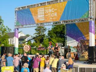 A live band performs on an outdoor stage at Grand Junction Plaza during the Jams at the Junction concert, with a crowd gathered in front on a sunny day.