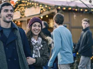 A smiling couple in winter attire enjoying the atmosphere of an outdoor holiday market with wooden stalls in the background.