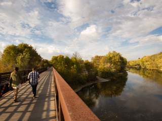 Two people walk along a pedestrian bridge at Forest Park beside a calm river, surrounded by trees under a partly cloudy sky.