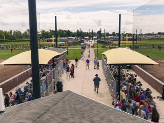 An elevated view of a wide central walkway with people walking between two long covered seating areas. Sports fields extend on both sides, with groups gathered under the shelters and open green space stretching into the distance.