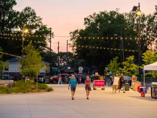 People walk through Grand Junction Plaza at dusk during the June 2023 Jams at the Junction event, with string lights glowing above vendor tents and trees.