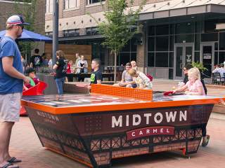 People gathered in a vibrant outdoor plaza around a large, bright orange ping-pong table labeled "Midtown Carmel."