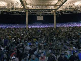 An outdoor stage venue with a crowd gathered in front of a stage featuring large screens, set beneath a steel-framed overhead canopy.