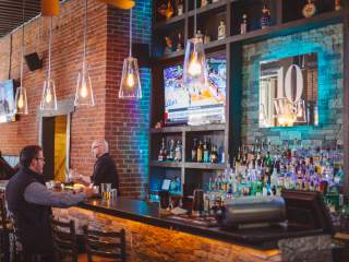 A bar interior with exposed brick walls, hanging pendant lights, a long bar counter, shelves of bottles, and a wall display showing the text ‘10 West,’ with people seated at the bar.