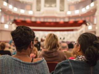 The backs of seated audience members fill the foreground, facing a brightly lit concert hall interior with a stage, balcony seating, and warm architectural lighting visible ahead.