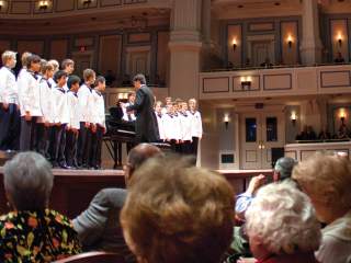 Low-angle view from the audience of a stage where performers in white jackets stand in rows, with a conductor in a black jacket facing them at the front. Audience members fill the seats in the foreground, and multi-level balconies rise along the back and sides of the venue.