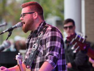 A person singing into a microphone on an outdoor stage, with another musician visible in the background.