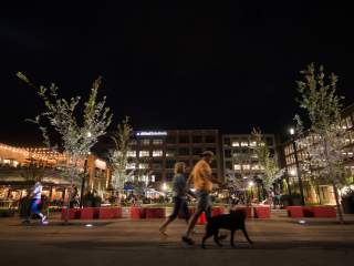 A lively Carmel Midtown Plaza at night, featuring illuminated trees, outdoor seating, and people walking along a pathway.