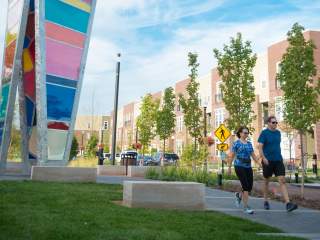 Two people jogging along a paved urban trail beside a tall, colorful glass art structure, with trees, benches, and multi-story residential buildings lining the path.