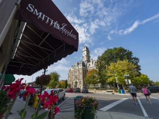 A streetscape view with a brick building featuring a clock tower in the background, a sidewalk lined with flowers and parked cars, and an awning with a business name partially visible in the foreground.