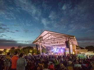 A large crowd gathered outdoors at night facing a covered stage with bright lights, with people standing closely together in front of the stage under a darkening sky.