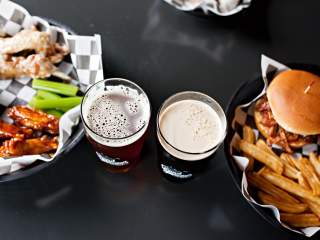 A top-down view of two glasses of dark beer sitting on a black table alongside baskets of wings, a sandwich, and french fries.