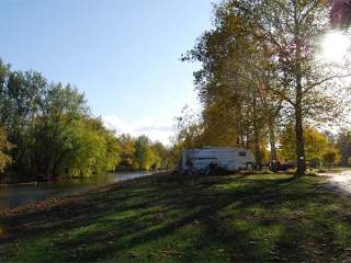 A scenic view of a calm river reflecting the green trees on its banks under a clear blue sky, and RV camper is in the background.