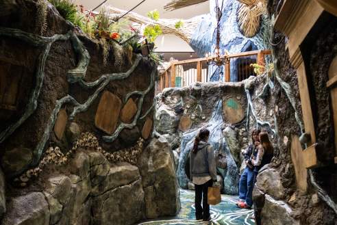 Two visitors explore a whimsical, nature-inspired indoor pathway with rocky textures, greenery, and artistic details. The scene invites adventure.
