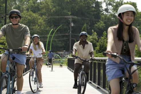 A group of friends ride bicycles across a wooden bridge at Grand Junction Plaza, smiling as they follow the trail surrounded by greenery.
