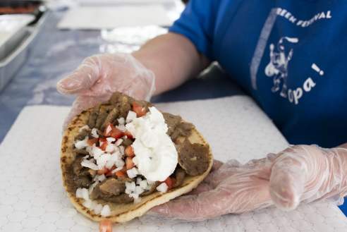 A freshly prepared gyro on warm pita bread is held by a food service worker wearing clear gloves. The sandwich is topped with sliced meat, diced onions, chopped tomatoes, and a generous dollop of creamy tzatziki sauce. The worker wears a blue shirt, and the gyro is presented over deli paper at a restaurant counter.