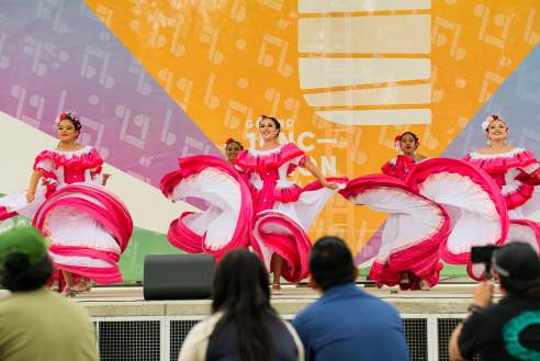 Dancers in bright red traditional dresses perform a folkloric dance on an outdoor stage at the Cuisine Connect Culture event, with spectators watching from the audience.