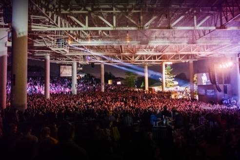 Large crowd gathered at an outdoor concert venue at night, facing a brightly lit stage beneath a covered pavilion structure. Overhead beams and lighting rigs frame the scene, with the audience filling the foreground and middle ground.
