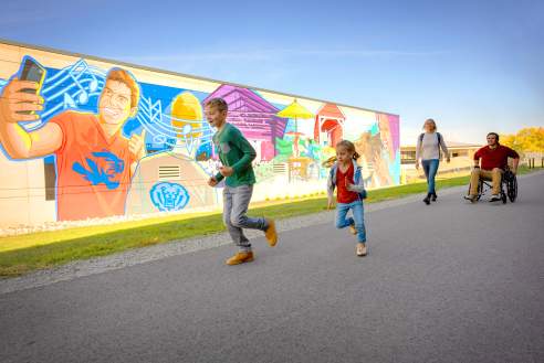 A group of people moving along a flat paved trail beside a long, colorful mural painted on a wall, with one person using a wheelchair on the right and others walking or running ahead, while the mural features large, brightly colored figures and graphic patterns.