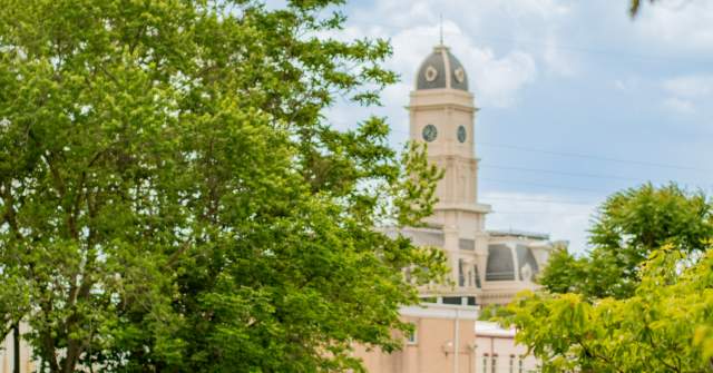 Historic courthouse building surrounded by trees and greenery, photographed in warm daylight. People ride rail bikes in the foreground.