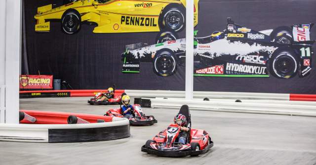 Three people drive red go-karts around an indoor track at K1 Speed, with large racing car murals on the wall behind them.