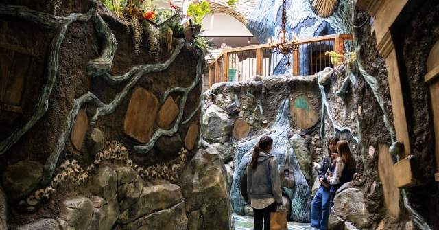 Two visitors explore a whimsical, nature-inspired indoor pathway with rocky textures, greenery, and artistic details. The scene invites adventure.