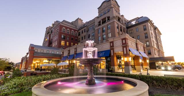 A mixed-use urban complex at dusk with multi-story brick and stone buildings lit from within. In the foreground, a small fountain with water flowing into a basin sits in a landscaped plaza, while storefronts and sidewalks line the street.