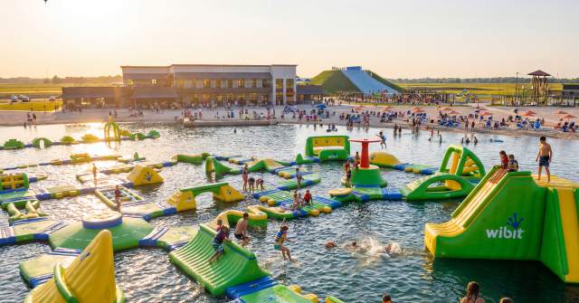 A vibrant water park scene featuring inflatable structures in a lake, with families enjoying activities under a sunset sky. A beach area is visible.