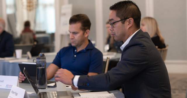 Two men sit at a conference table at Hotel Carmichael, working on laptops and tablets during a meeting session.