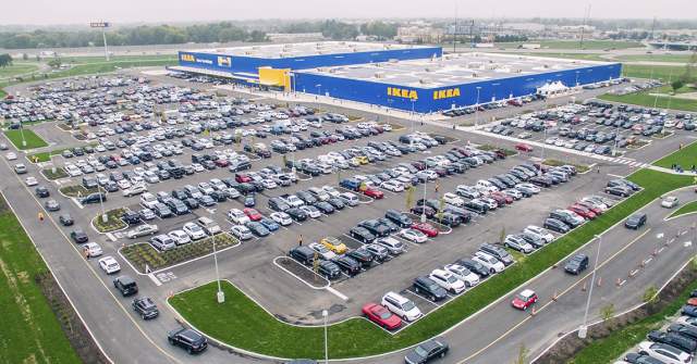 A high-angle view of a parking lot filled with cars, bordered by green landscaped areas and wrapping around a large blue, warehouse-style building with yellow accents.