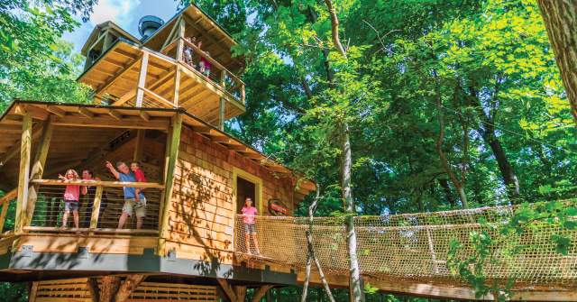 A multi-story wooden treehouse structure built among tall green trees with children playing.