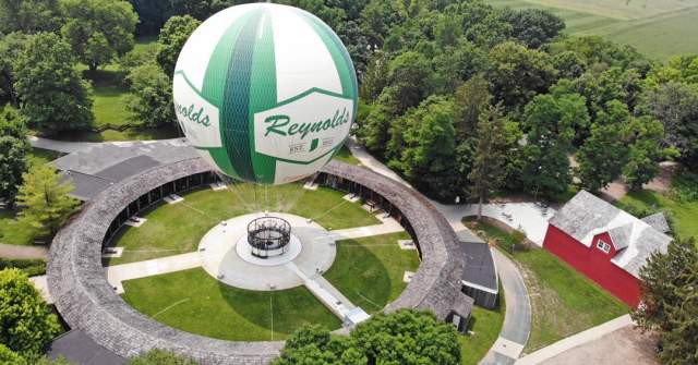Hot air balloon in vibrant green and white colors floating above a grassy field, trees, and a barn below at Conner Prairie.