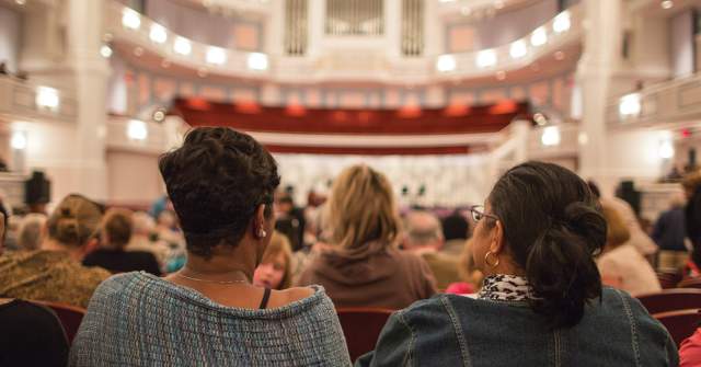 The backs of seated audience members fill the foreground, facing a brightly lit concert hall interior with a stage, balcony seating, and warm architectural lighting visible ahead.