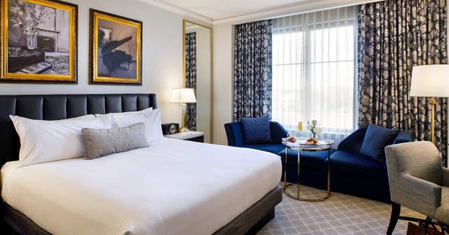 A hotel room with a king-size bed dressed in white bedding, a dark upholstered headboard, and framed artwork above the bed. The room includes a seating area with blue chairs, a small table, floor lamps, and patterned curtains framing a large window, creating a modern, upscale atmosphere.