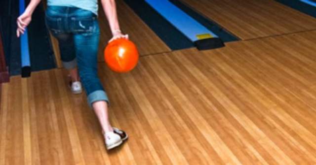 A child wearing a blue shirt and jeans rolling an orange bowling ball down a dimly lit lane with light orange wooden floors.