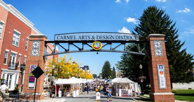 Decorative street arch reading “Carmel Arts & Design District” above a pedestrian street lined with white vendor tents, trees, and storefronts.