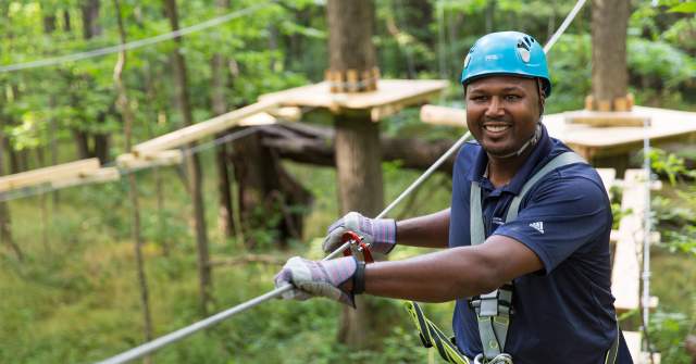 A person wearing a blue safety helmet and harness smiles while navigating an aerial ropes course among the trees.