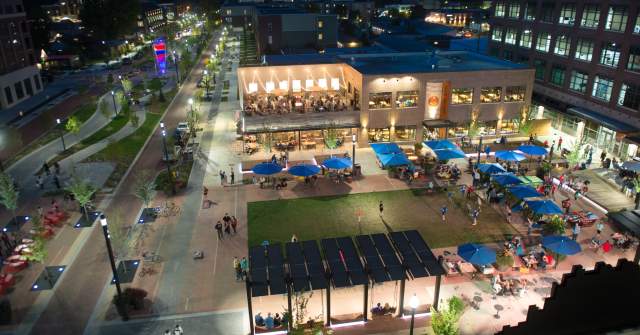 A nighttime aerial view of Midtown Plaza shows a lively, illuminated urban gathering space surrounded by buildings. The plaza features open green areas, walkways, and multiple outdoor seating zones with blue umbrellas, where people gather and dine. Streetlights, storefront lighting, and passing traffic line the adjacent streets, while nearby multi-story buildings frame the scene, conveying an active, vibrant downtown atmosphere after dark.