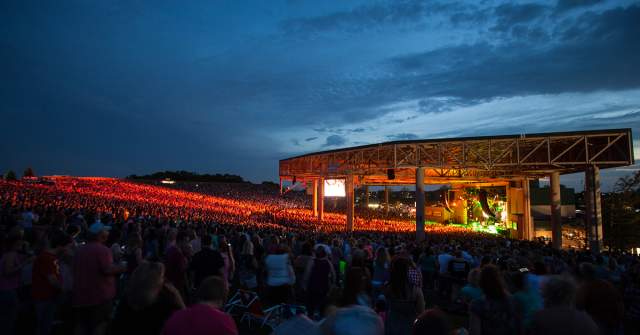 Large outdoor concert venue at dusk, with a brightly lit stage and a crowd filling the seating area. Rows of lights glow across the venue under a darkening blue sky.