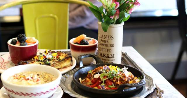 A tabletop display of breakfast dishes including baked eggs in small ramekins, a skillet topped with vegetables and herbs, a bowl of soup or grits, and a small vase of flowers, set on a café table with a yellow chair in the background.
