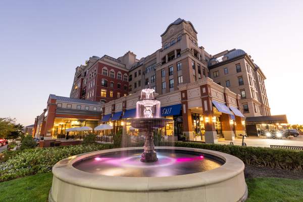 A mixed-use urban complex at dusk with multi-story brick and stone buildings lit from within. In the foreground, a small fountain with water flowing into a basin sits in a landscaped plaza, while storefronts and sidewalks line the street.