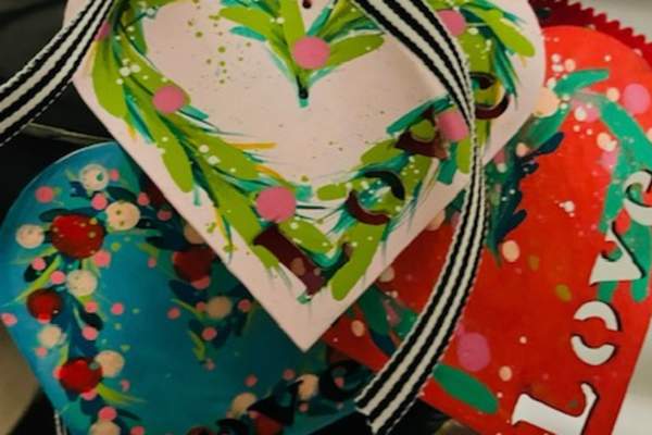 Several colorful, hand‑painted heart-shaped tags decorated with splattered dots and brushstroke designs hang together from striped and twine ribbons on a small black stand. The tags feature bright greens, pinks, blues, reds, and the word ‘Love’ on some of them.