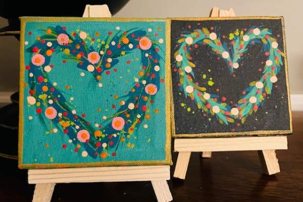Two small square paintings on miniature easels show colorful heart designs made of swirling brushstrokes and scattered dots—one on a teal background and the other on a dark background.