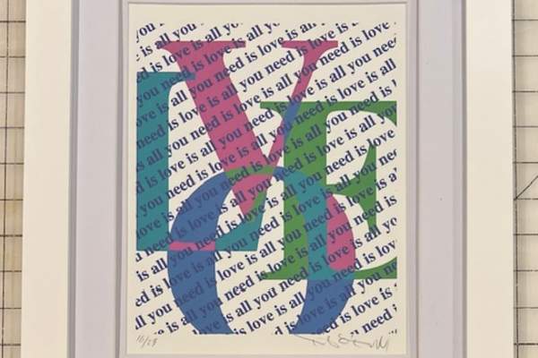 A framed print featuring large overlapping letters in blue, green, and pink that spell ‘LOVE,’ layered over a repeating diagonal background text that reads ‘all you need is love.’