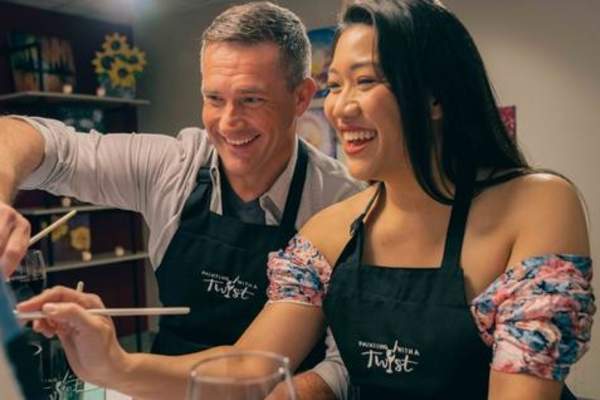 Two people wearing black ‘Painting with a Twist’ aprons sit side by side at a painting class, holding brushes and working on canvases in a warmly lit studio.
