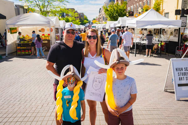 A family poses in the middle of a street lined with art vendor tents during the Carmel International Arts Festival. Two children wear balloon Viking helmets while crowds browse booths in the background.