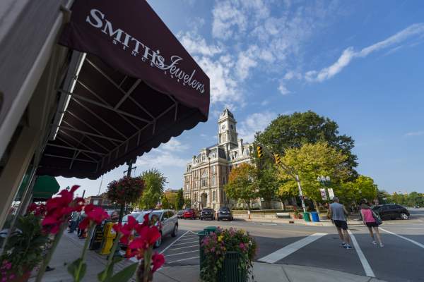 A streetscape view with a brick building featuring a clock tower in the background, a sidewalk lined with flowers and parked cars, and an awning with a business name partially visible in the foreground.